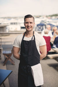 Portrait of smiling young waiter wearing apron on restaurant patio