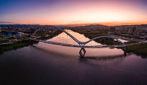 Bridge over river by buildings against sky during sunset