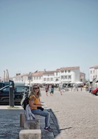 Woman sitting on bollard at sidewalk in city against clear sky