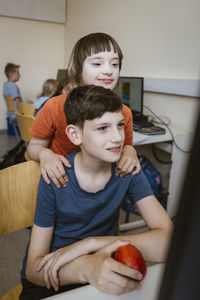 Girl with down syndrome near male friend sitting on chair in computer class at school