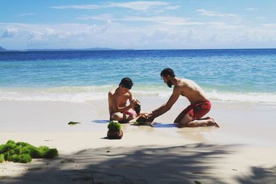 Shirtless men breaking seashell at beach against sky