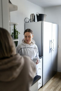 Smiling young woman holding coffee cup while standing near girlfriend in kitchen