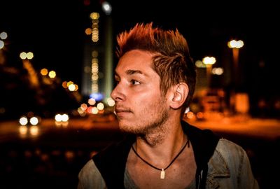 Close-up of man looking away against illuminated buildings at night