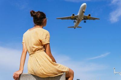 From below back view of unrecognizable female in casual clothes sitting against blue sky with plane flying in blue sky