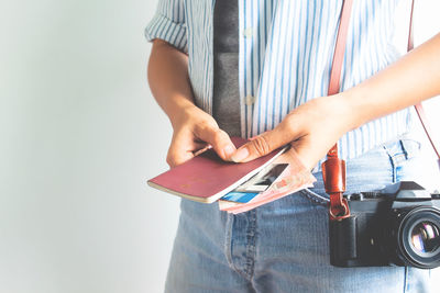 Midsection of woman holding passport with paper currency and camera standing against white background