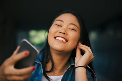 Cheerful teenage girl adjusting in-ear headphones while holding smart phone in city