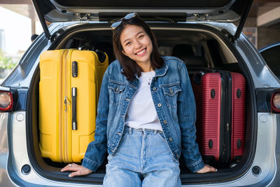 Happy asian young woman sit on suv car trunk with suitcases or luggages. she ready holiday vacation