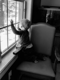 Baby boy kneeling on chair while looking through window at home