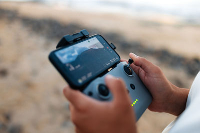 Soft focus of unrecognizable ethnic tourist using remote control and smartphone to fly drone over sea and shoot waves on weekend day on beach