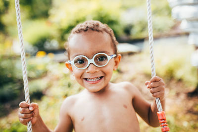 Portrait of shirtless boy sitting on swing against trees