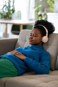 Resting african american woman in headphones with slight smile lies on sofa falling asleep to music