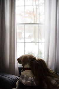 Rear view of girl embracing dog while sitting by window
