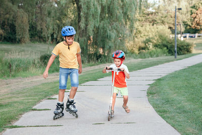 Boy skating while brother riding push scooters on footpath in park