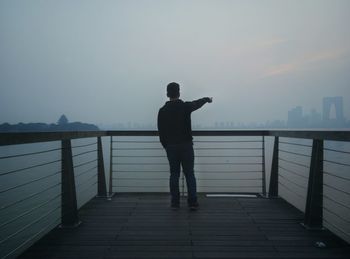 Man standing on railing by sea against sky during sunset