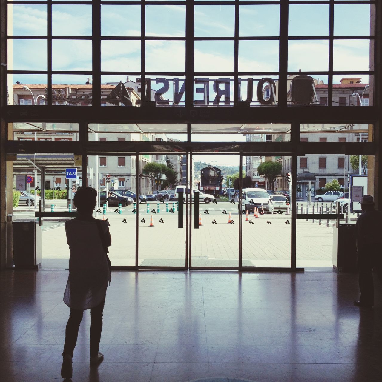 indoors, glass - material, men, window, lifestyles, architecture, person, transparent, built structure, walking, rear view, railroad station, city, leisure activity, full length, standing, city life, reflection, public transportation