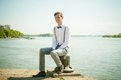 Portrait of smiling young man sitting on bollard against river during sunny day