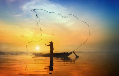 Silhouette fisherman standing on boat while throwing fishing net in sea against sky during sunrise