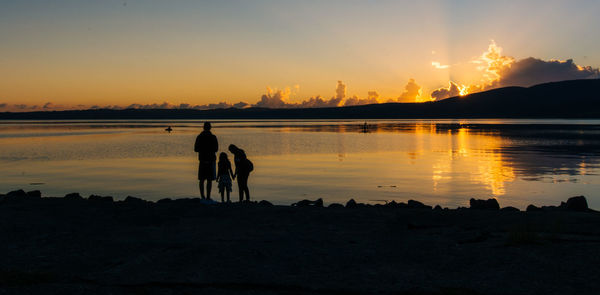 Silhouette family on lakeshore against sky during sunset