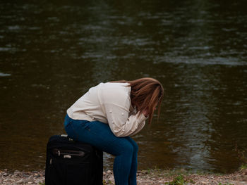 Woman sitting on luggage against lake