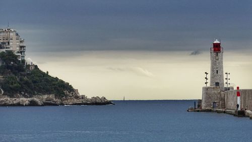 Lighthouse by sea and buildings against sky