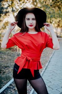 Portrait of beautiful young woman wearing hat standing on footpath against trees