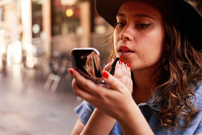 Young female in hat looking in mirror and smearing red lipstick on lips while applying makeup on street of madrid, spain