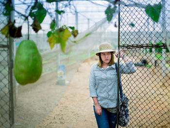 Young woman looking away while standing by fence at farm