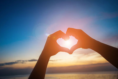 Close-up of hand making heart shape against sky during sunset