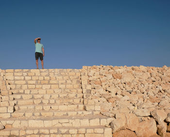 Low angle view of man shielding eyes while standing on steps against clear blue sky