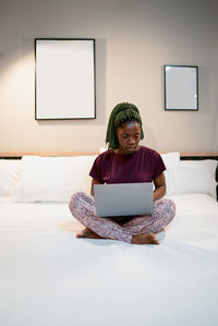 Full body of young ethnic female sitting on bed and browsing netbook while working remotely in modern bedroom at home