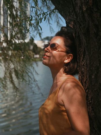 Side view of smiling young woman wearing sunglasses standing by tree