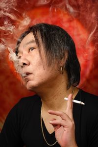 A man in black smoking a cigarette in front of a red background, blowing smoke out of his mouth.