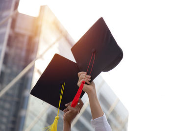 Low angle view of people hands holding mortarboards against sky