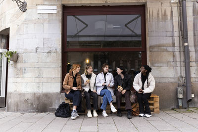 Young happy female friends talking to each other while sitting on pallets at sidewalk in city