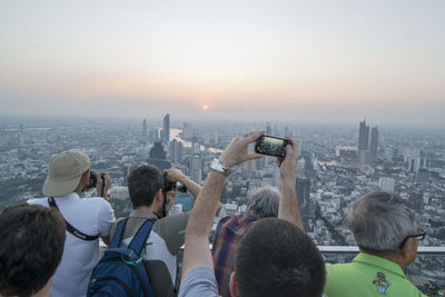Rear view of people photographing cityscape against sky during sunset