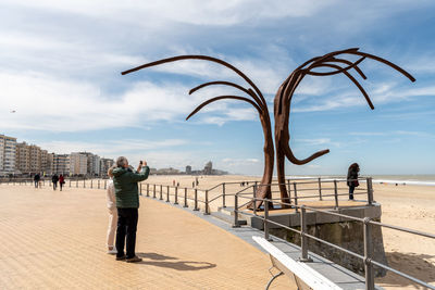 Couple photographing sculpture on promenade in city against sky