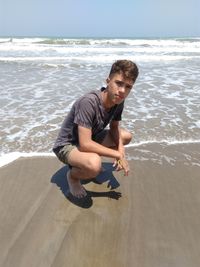 Portrait of teenage boy crouching on sand against sea at beach