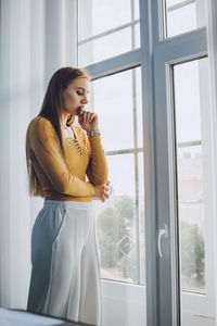 Young woman looking away while standing on window
