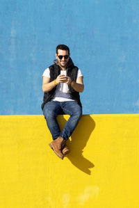 Young bearded man with sunglasses sitting on yellow wall, using phone