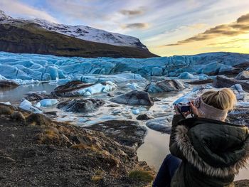 Rear view of mid adult woman photographing frozen sea while sitting at beach against sky during sunset