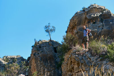 Low angle view of young man standing on rock against clear blue sky