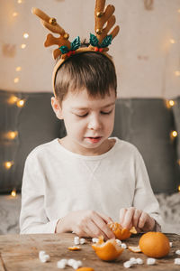 Boy in christmas antlers, sits and peels a tangerine on the background of a yellow garland.