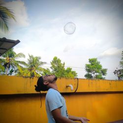 Side view of young man playing with bubble against sky