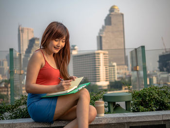 Beautiful young woman writing in book while sitting in city