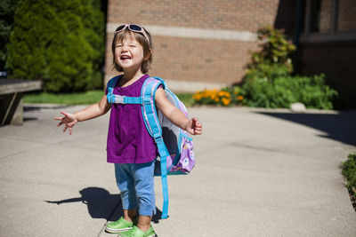 Portrait of happy baby girl with backpack standing on footpath during sunny day