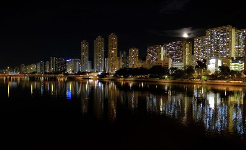 Illuminated buildings by river against sky at night