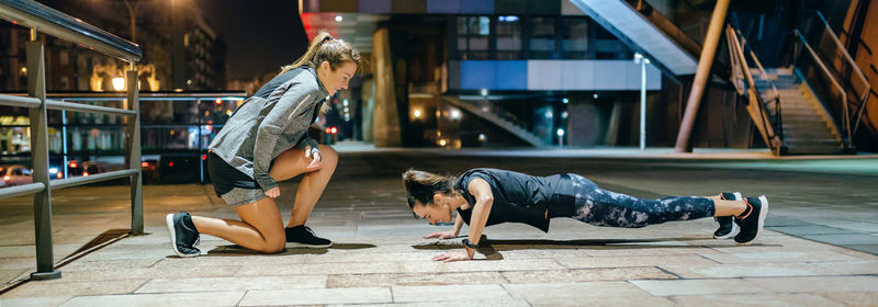 Woman doing push ups by supervision of personal trainer on the city at night