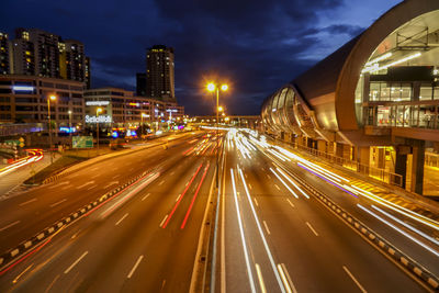 Light trails on road by illuminated buildings against sky at night