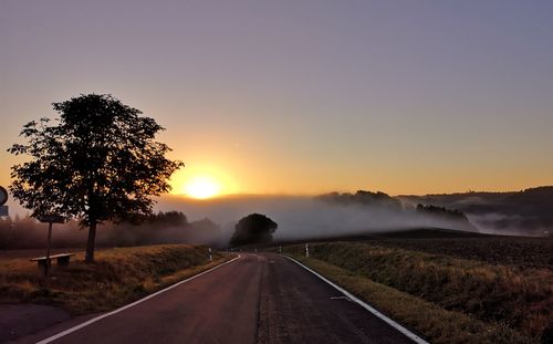 Road by trees against sky during sunset