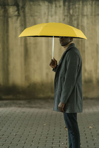 Young man with yellow umbrella staring while standing on footpath